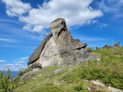 A large, uniquely shaped rock formation on a grassy hillside under a blue sky with scattered clouds.