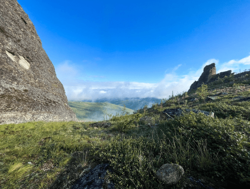 A panoramic view of a rocky landscape with green hills and a blue sky, partially covered by mist.