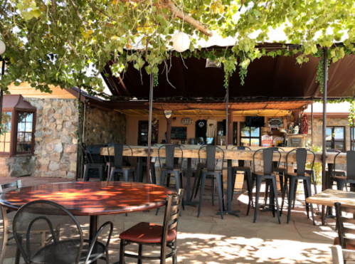 Outdoor seating area of a rustic bar with stone walls and a wooden counter, shaded by leafy vines.