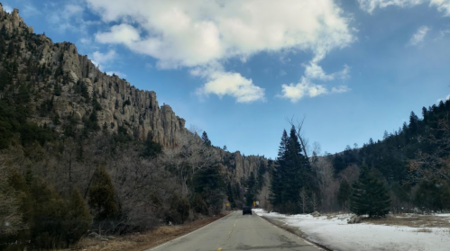 A scenic road winds through rocky cliffs and trees under a partly cloudy sky, with patches of snow along the roadside.