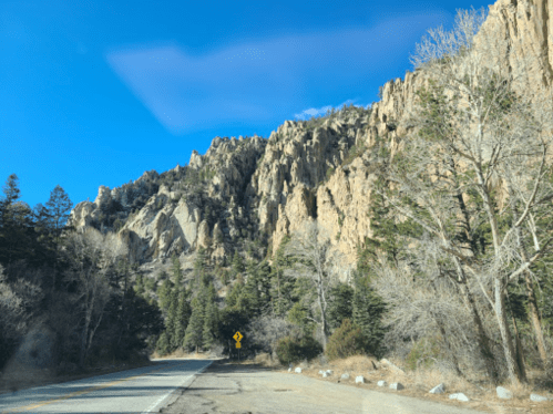 A scenic view of rocky cliffs and trees alongside a winding road under a clear blue sky.