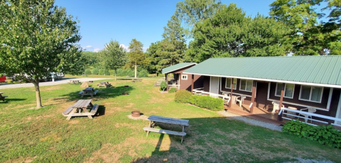 A scenic view of a cabin area with green grass, trees, picnic tables, and a clear blue sky.