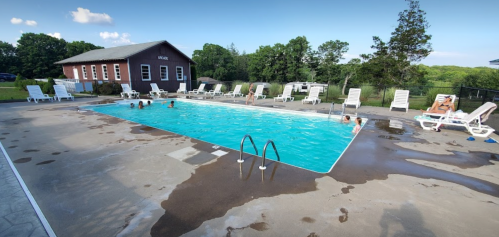 A sunny outdoor pool surrounded by lounge chairs, with people swimming and relaxing nearby. A building is in the background.