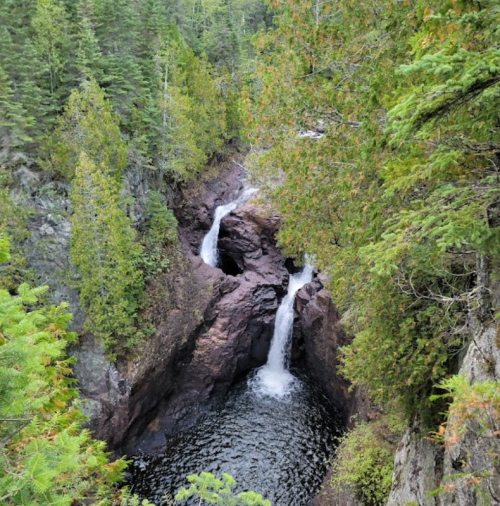 A scenic view of a waterfall cascading into a dark pool, surrounded by lush green trees and rocky cliffs.