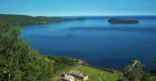 A serene view of a blue lake with an island, surrounded by green trees and a historic building in the foreground.