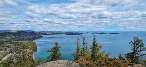 A panoramic view of a serene lake surrounded by lush greenery and distant hills under a partly cloudy sky.