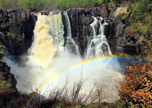 A powerful waterfall cascades over rocks, creating a misty spray with a vibrant rainbow arcing across the scene.