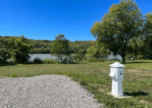 A white post with the number 54 stands near a river, surrounded by green trees and a clear blue sky.