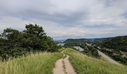 A winding dirt path leads through tall grass, overlooking a river and hills under a cloudy sky.