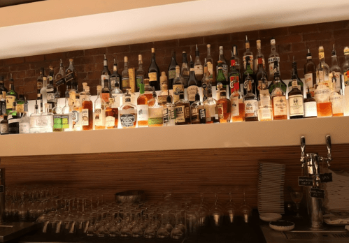 A well-stocked bar shelf displaying various bottles of liquor against a brick wall, with glasses in the foreground.