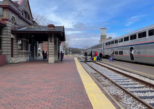 A train station with passengers boarding a train, brick pathway, and cloudy sky in the background.