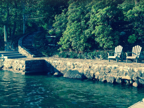 Two wooden chairs sit on a stone patio by a calm lake, surrounded by lush greenery and a winding path.