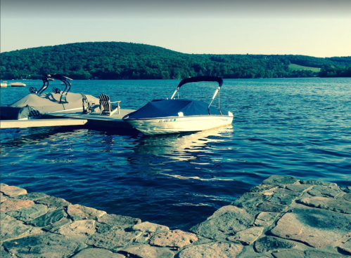 A boat docked by a stone pier on a calm lake, surrounded by green hills under a clear sky.