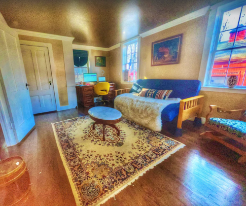 Cozy room with a blue sofa, wooden desk, and a patterned rug, featuring warm lighting and large windows.