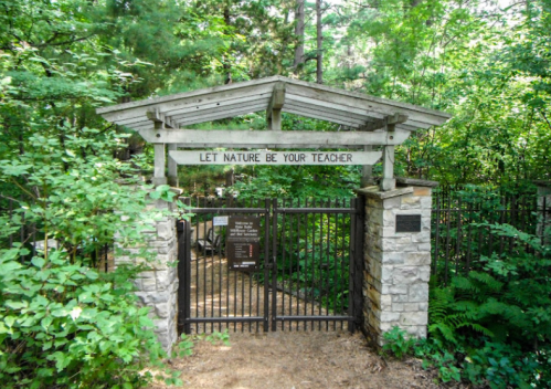 A wooden gate with a sign reading "Let Nature Be Your Teacher," surrounded by lush greenery and trees.