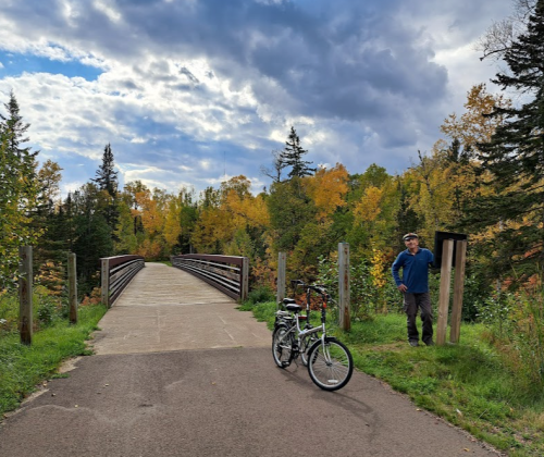 A cyclist stands beside a bike on a path leading to a wooden bridge, surrounded by trees with autumn foliage.