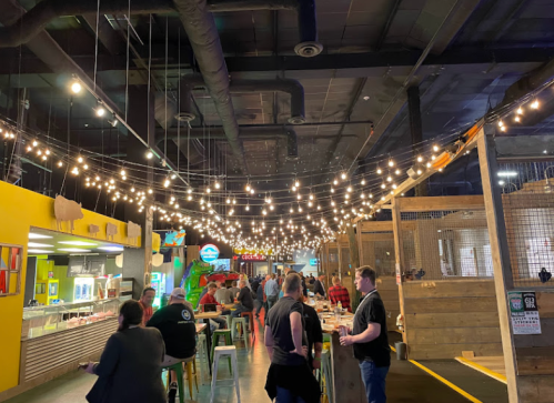 A lively indoor space with string lights, people dining, and a colorful food counter in a casual atmosphere.
