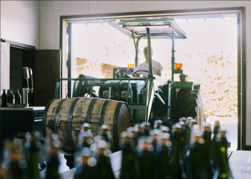 A tractor drives into a winery, with barrels and bottles in the foreground, and a worker visible in the background.
