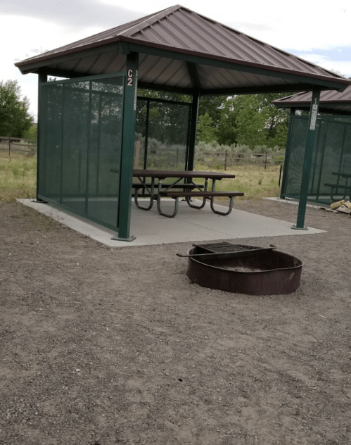 A picnic shelter with a table and a fire pit in a grassy area, surrounded by trees and open space.