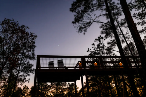A wooden deck surrounded by trees at dusk, with two chairs and a crescent moon in the sky.