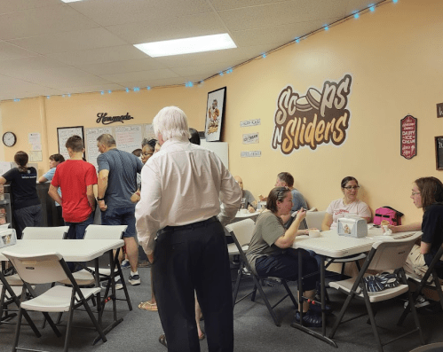 A busy ice cream shop with people socializing at tables and a wall featuring the "Scoops N Sliders" sign.