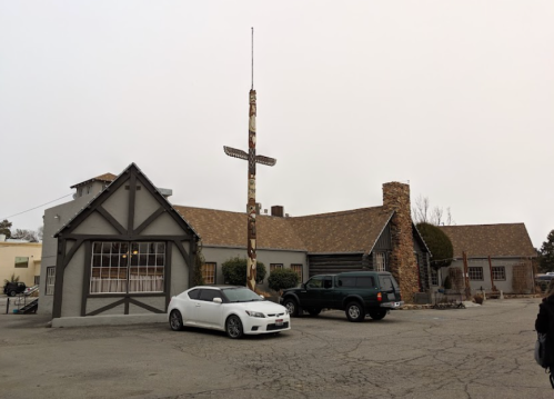 A gray building with a stone chimney and a totem pole in front, featuring parked cars in the lot.