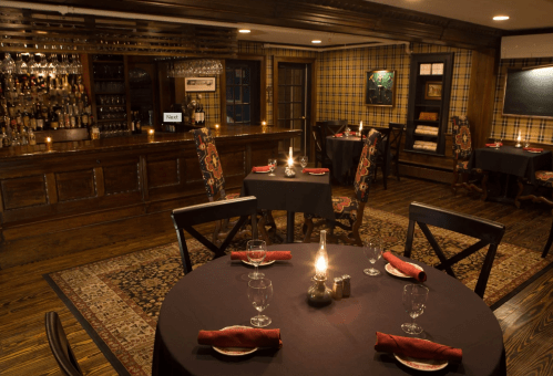 Cozy restaurant interior with dark tables, candlelight, and a wooden bar, featuring plaid walls and elegant decor.