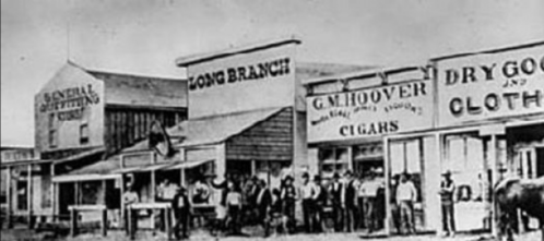 Historic black-and-white photo of a Western town with storefronts and a group of people gathered outside.