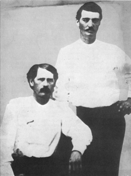 Two men in white shirts, one seated and one standing, posing for a vintage photograph.