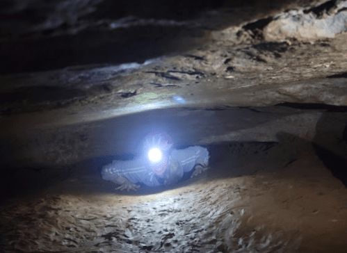 A person crawls through a narrow cave passage, illuminated by a headlamp, surrounded by rocky walls.