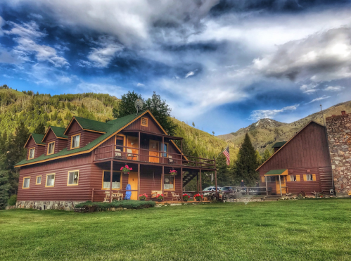 A rustic wooden lodge surrounded by mountains and greenery, with a blue sky and clouds overhead.