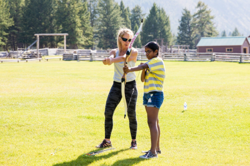 A woman teaches a girl how to use a bow and arrow on a sunny day in a grassy field surrounded by trees.