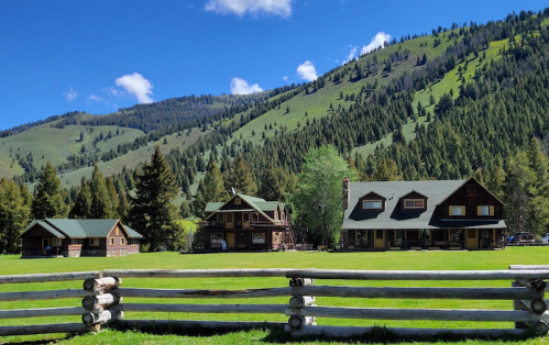 A scenic view of three rustic cabins surrounded by lush green hills and trees under a bright blue sky.