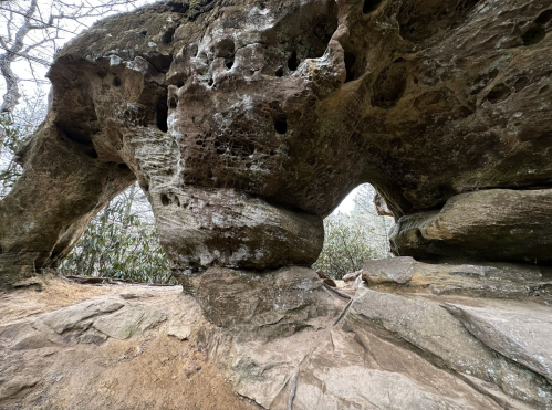 A large, weathered rock formation with a natural arch, surrounded by sparse vegetation and a cloudy sky.