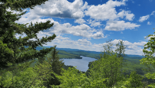 A panoramic view of a serene lake surrounded by lush green trees and rolling hills under a partly cloudy sky.