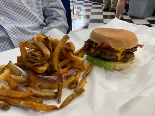 A close-up of a cheeseburger with bacon and lettuce, served with a side of golden French fries on a white table.