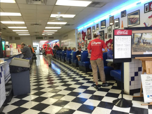 A diner with checkered floors, blue booths, and patrons enjoying meals, while staff in red shirts serve customers.