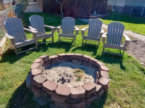 A circular stone fire pit surrounded by five gray Adirondack chairs on a grassy area.