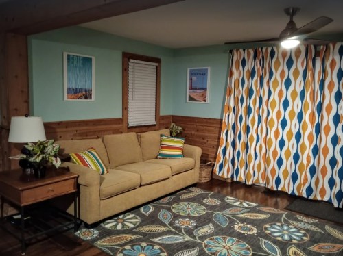 Cozy living room with a beige sofa, colorful curtains, and decorative rug, featuring wall art and a lamp.