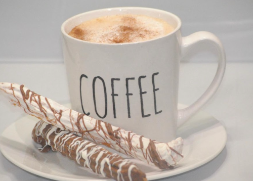 A white mug labeled "COFFEE" filled with frothy coffee, accompanied by two chocolate-drizzled snacks on a plate.