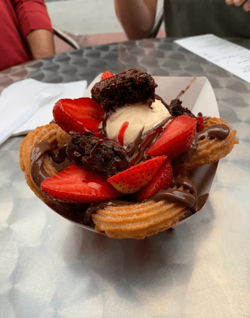 A dessert bowl featuring churros topped with strawberries, brownie pieces, chocolate sauce, and a scoop of ice cream.