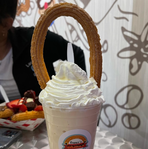 A creamy milkshake topped with whipped cream and a large churro shaped like a ring.