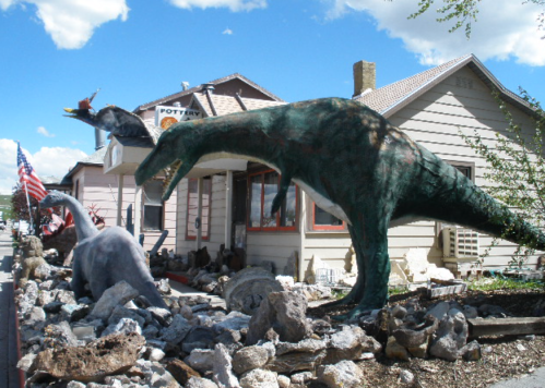 Colorful dinosaur sculptures in front of a pottery shop, with a blue sky and clouds in the background.