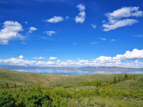 A scenic landscape featuring green hills, a calm lake, and a bright blue sky with fluffy white clouds.