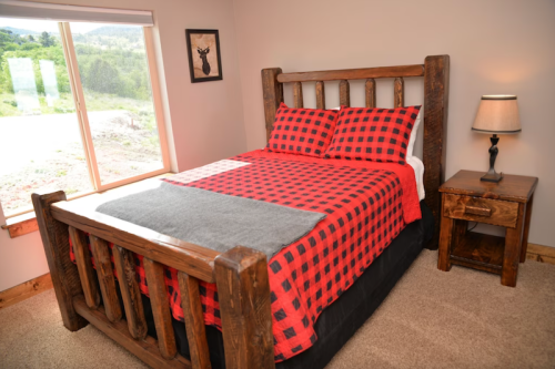 Cozy bedroom featuring a wooden bed with a red and black checkered blanket, nightstand, and large window with a view.