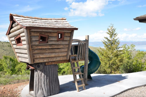 A wooden treehouse play structure with a slide and ladder, set against a scenic outdoor backdrop.