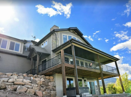 A modern house with a large deck, stone foundation, and blue sky in the background. Sunlight shines on the exterior.