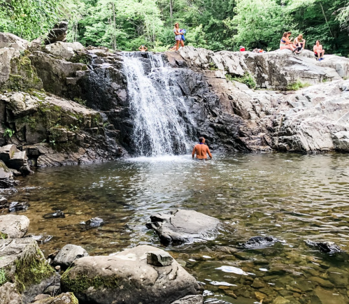 A small waterfall cascades into a rocky pool, with people relaxing on the rocks nearby in a lush green setting.