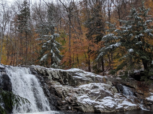 A serene waterfall cascades over rocky terrain, surrounded by trees with autumn leaves and a light dusting of snow.