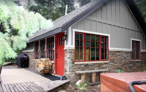 A cozy cabin with a red door, stone accents, and large windows, surrounded by trees and a wooden deck.